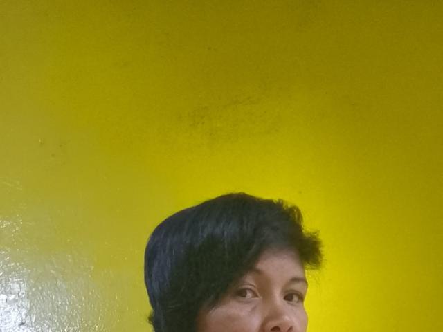 AnnaRosa69's face, upper chest, with short black hair, yellow wall behind, small dark spot near top of wall, slightly visible nose, and subtle smile.
