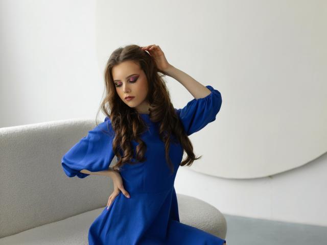 MileyGloss with long brown wavy hair, wearing a blue dress, sits on a gray sofa, hand in hair, other hand on hip, purple eye makeup, looking down, seductive.