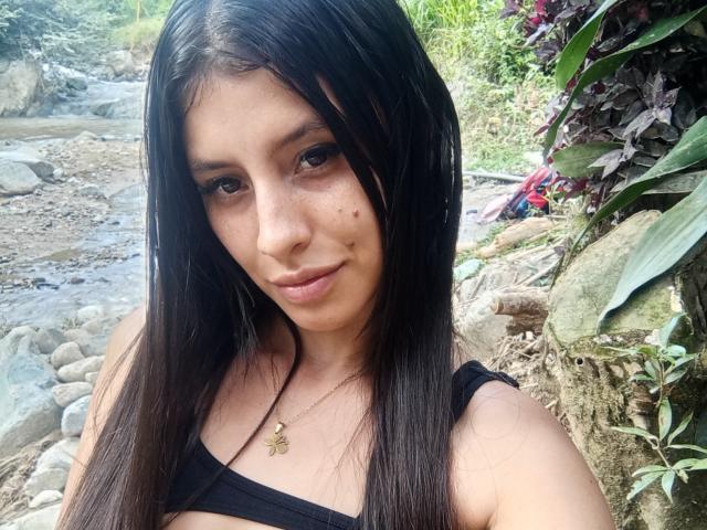 MadissonFontaine has long black hair, brown eyes, and a small nose with freckles. She's wearing a black top, showing her tits, and a butterfly necklace, smiling at the camera.