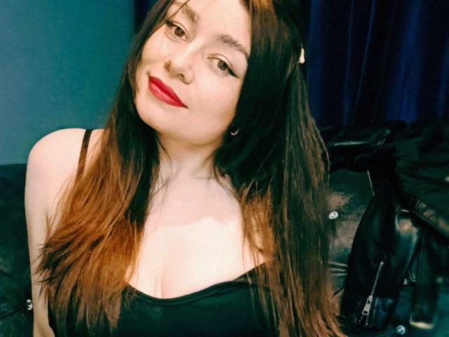 Michonnek with long brown hair, red lipstick, black tank top, smiling, fair skin, small tits, black leather jacket on couch, blue curtain in background.
