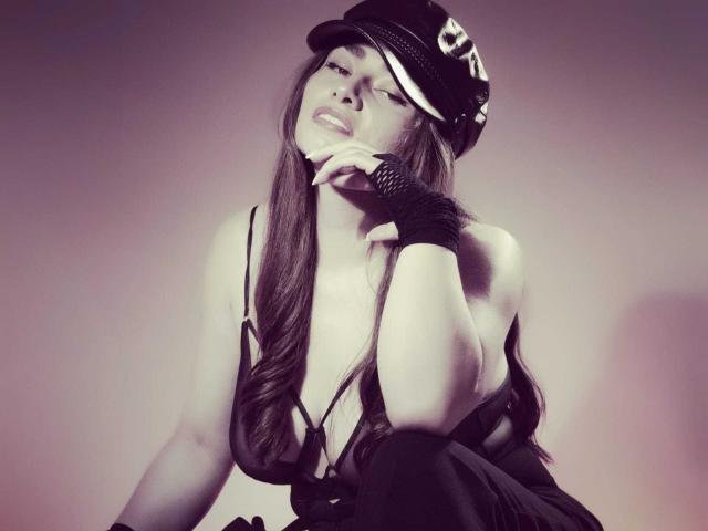 Michonnek with long brown hair, black hat, and fingerless glove, leaning forward, showing cleavage, looking seductive, with a slight smile, in a monochrome, slightly blurry photo.