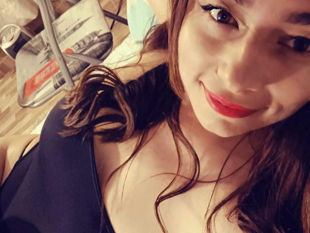 Michonnek has medium tits, a cute face with red lipstick, brown wavy hair, and a black top, smiling at the camera. Her ass is partially visible.
