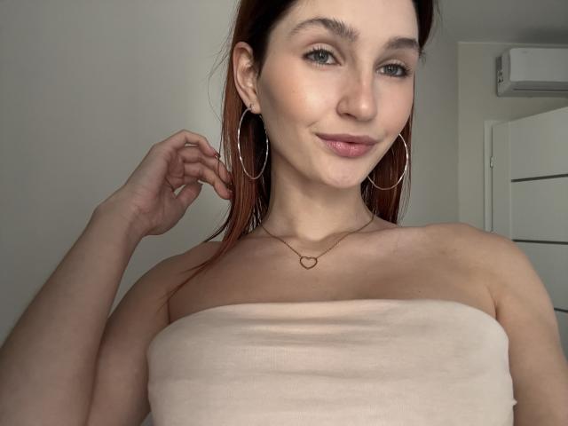 CarmelinaLove with brown hair, green eyes, and hoop earrings, wearing a strapless beige top, smiling, hand near ear, heart necklace, pale skin, slightly flushed, in a selfie.