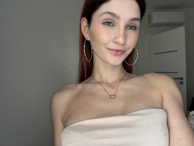 CarmelinaLove with pale skin, blue eyes, and brown hair, wearing hoop earrings and a heart necklace, smiling at the camera, topless with her tits covered by a white cloth.