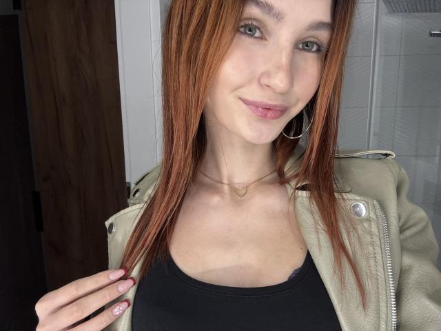 CarmelinaLove has red hair, green eyes, and a small smile. She's wearing a green jacket over a black top, with a heart necklace. Her hand's on her chest.