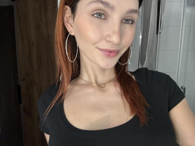 CarmelinaLove with light skin, green eyes, and reddish-brown hair, wearing a black tee, hoop earrings, and a heart necklace, smiling slightly with pink lipstick.