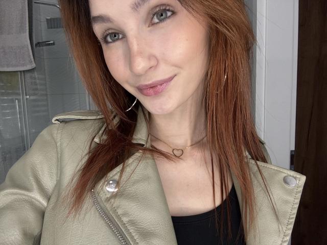 CarmelinaLove with light skin, green eyes, and red hair, wearing a beige leather jacket and black top, small heart necklace, subtle smile, and small hoop earrings.