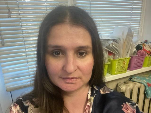 MegaBoom has medium-length brown hair, a slightly puffy face, and is wearing a floral shirt, standing in front of white blinds with a yellow basket of papers.