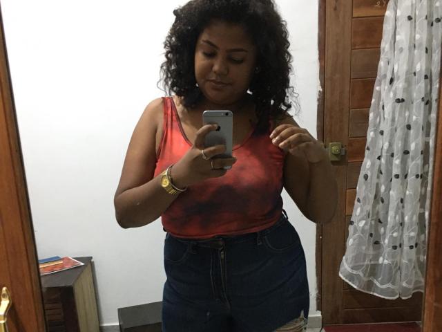 WendyStar69 with curly black hair, wearing a red tank top and high-waisted blue jeans, taking a mirror selfie. She has a gold watch and is looking down.