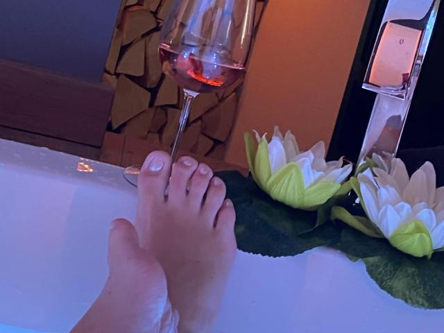 MisstressCathy-hot's feet in a white bathtub with pink wine in a glass, green lily pads, and white lotus flowers, under soft orange lighting.