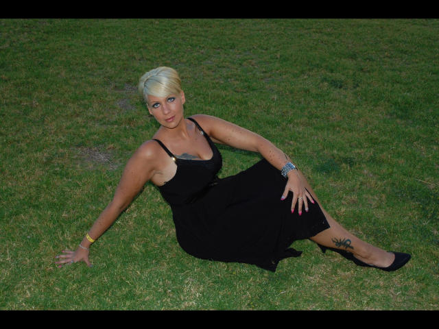 MistressCathy-hot with short blonde hair, black dress, and tattoos, sitting on grass with one arm behind her and the other on her knee, showing her ass and tits.