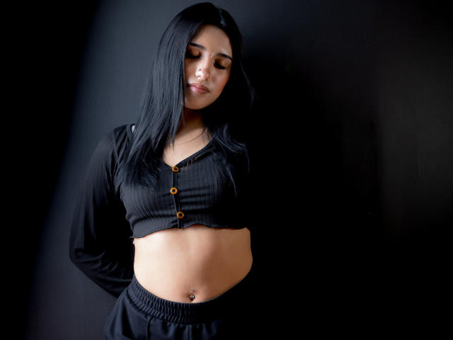 Nicki Clark, com cabelos longos e pretos, vestindo uma cropped preta com textura em tiras e botões, e calças pretas, exibindo um piercing no umbigo, em pé contra um fundo escuro..