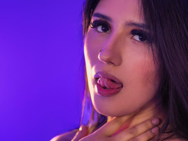 NickiClark has dark hair, a nose ring, and is sticking out her tongue with her hand on her neck. Her purple-lit face is sexy and she's looking at the camera.