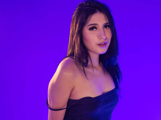 Nicki Clark, con cabello largo y oscuro, vestida con una blusa negra con un hombro descubierto, tiene un rostro adorable, un maquillaje sutil y una ligera sonrisa, sobre un fondo morado. Se pueden ver los senos..