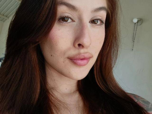 Isabella Palacio tiene ojos marrones, labios rosados y cabello castaño con una pequeña peca debajo del ojo derecho. Está mirando directamente a la cámara con una expresión neutra..