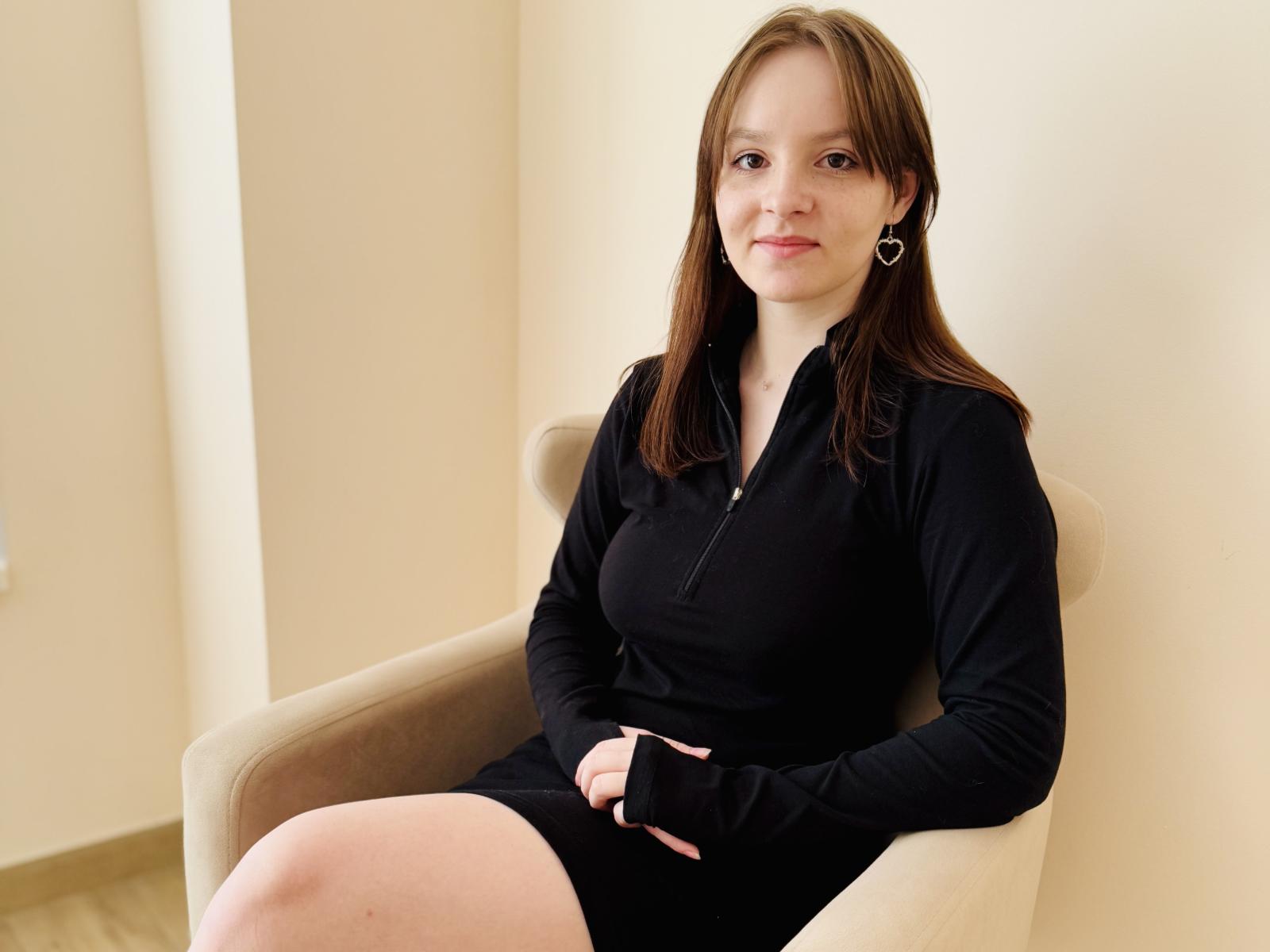 NancyMitford with brown hair, black zip-up dress, heart earrings, sitting in beige chair, smiling, hands on lap, visible ass, pale skin, light wood floor, casual setting.