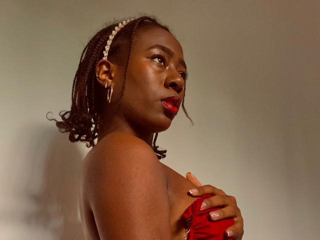 VioletGraice with dark skin, red lipstick, and braided hair wearing a red strapless top, covering her tits with her hand, looking to the side with a serious expression.