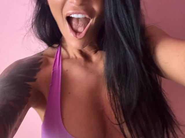 AndraD with long black hair, purple swimsuit, tongue out, showing teeth, tattoo on right shoulder, pink background, visible collarbone, and tan skin. Boobs, tongue, tattoo.