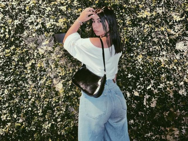 LanaYourDream with medium-ass in high-waisted jeans, white off-shoulder top, black shiny purse, and hand in hair, standing against a leafy wall. She has dark hair and a bracelet.