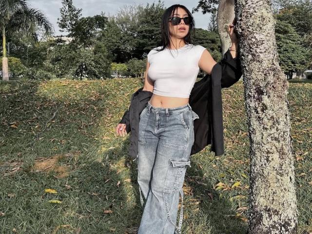 LanaYourDream with black hair, sunglasses, white crop top, and blue jeans, standing by a tree, black jacket in hand, showing a flat stomach and a small ass.