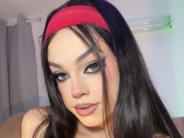 EvaaMooree has black hair, a red headband, and is staring with blue eyes, full lips, and thick black eyeliner. Her tits are not visible in the image.