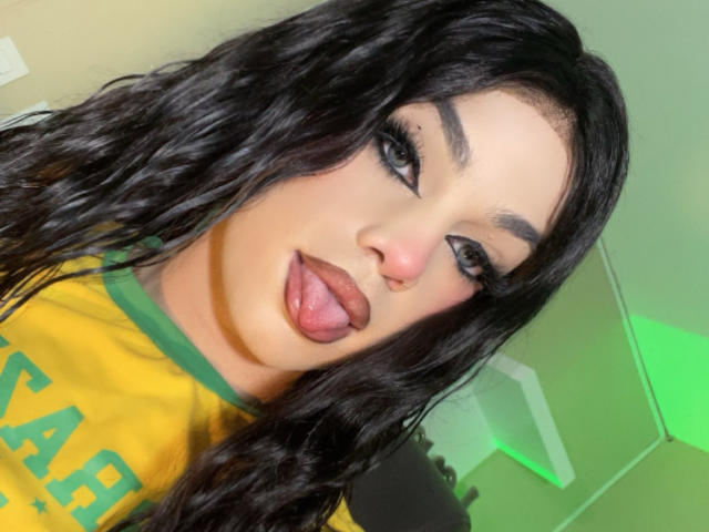 EvaaMooree with wavy black hair, sticking out her tongue, green and yellow shirt, and bold black eyeliner. Her nose is pierced on the right side.