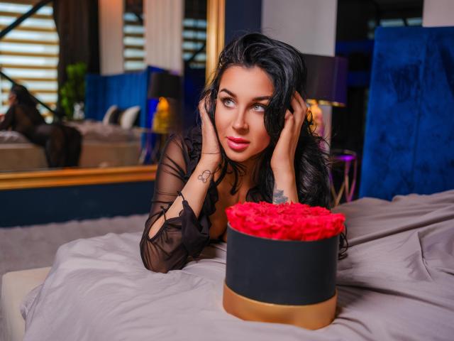FrancescaDante with black hair, blue eyes, and pink lipstick, wearing a sheer black top, hands on her face, looking at a red rose-covered black and gold box on a bed.