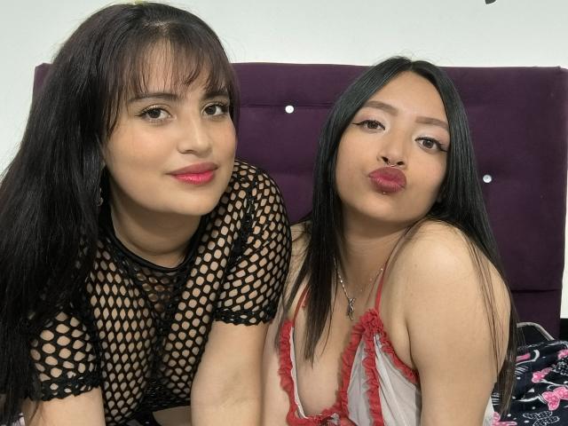 Two LesbianHotSl women with black hair, wearing fishnet and red lace lingerie, pouting their lips. One has medium breasts, the other has small tits, sitting in front of a purple headboard.