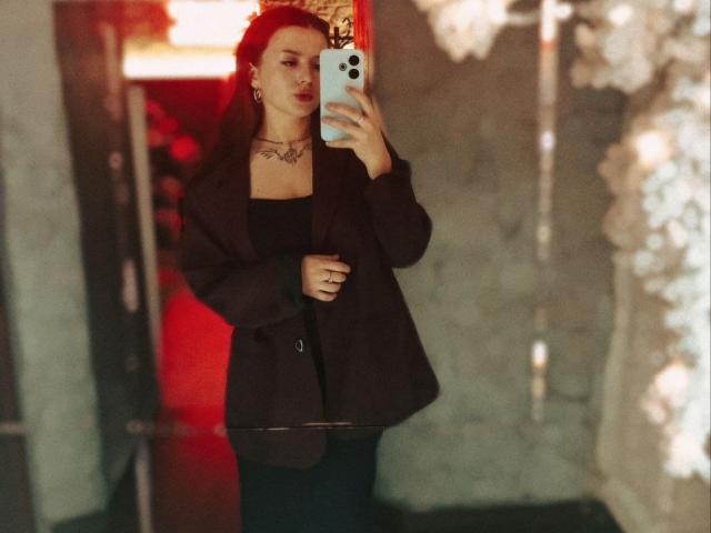 DaizyRara with dark hair, wearing a black top and dark jacket, holding a phone, taking a mirror selfie, showing a tattoo on her chest, in a dimly lit room.