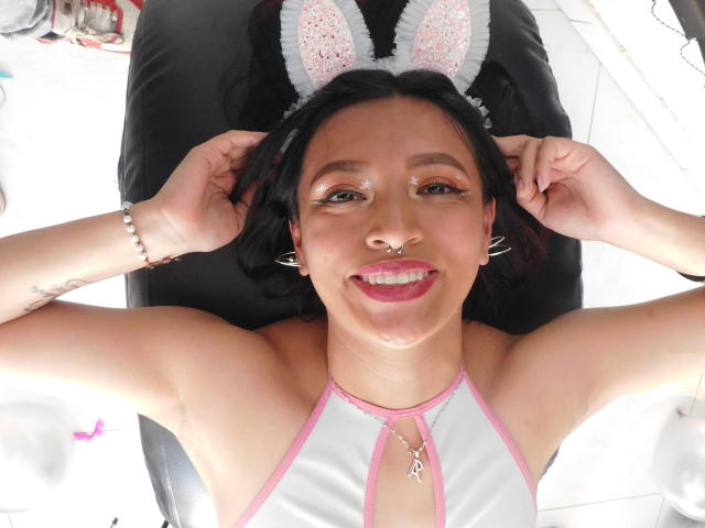 LesbianHotSl with cat ears, smiling, nose piercing, red lipstick, white and pink halter top, bracelet, necklace, lying on black chair, hands behind head, shiny face, small tits.