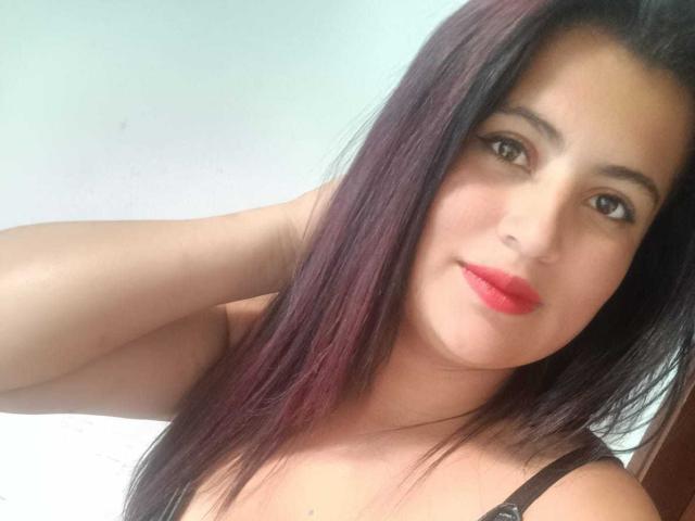 LesbianHotSl has long dark purple hair, red lipstick, brown eyes, and a slight smile, wearing a black strap, with her hand behind her head in a selfie.