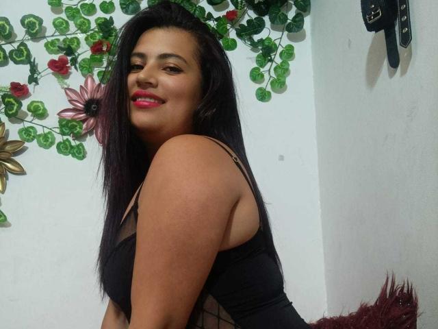 LesbianHotSl with long black hair, tan skin, pink lipstick, black top, smiling, showing her ass, green leaf decorations, and pink flower wall art in the background.