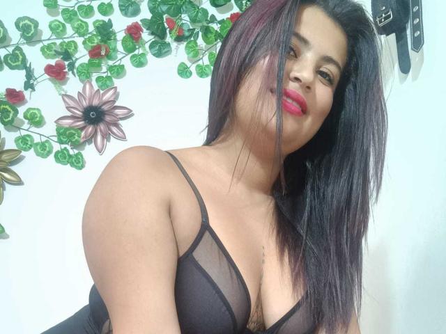 LesbianHotSl with medium brown skin and red lipstick, wearing a sheer black bralette, showing her tits and long black hair, smiling slightly. Her eyes are half-open.