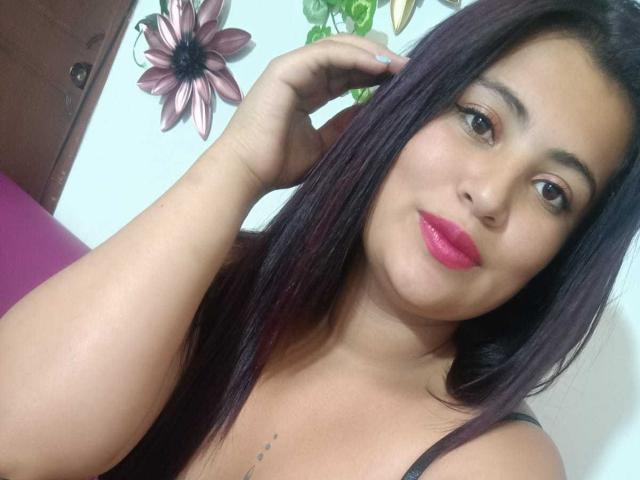 LesbianHotSl with dark purple hair, pink lipstick, and tan skin, touching her head, showing a tattoo on her chest, wearing a black top, smiling slightly.