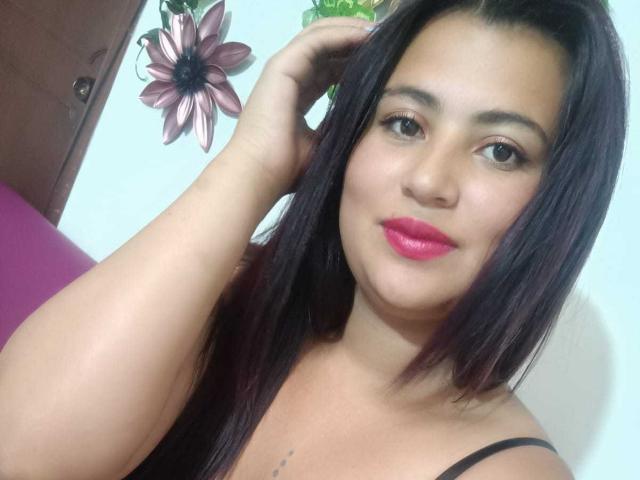 LesbianHotSl with dark brown hair, pink lipstick, and medium tits, touching her head with a hand, smiling slightly, wearing a black strap top, against a white wall.