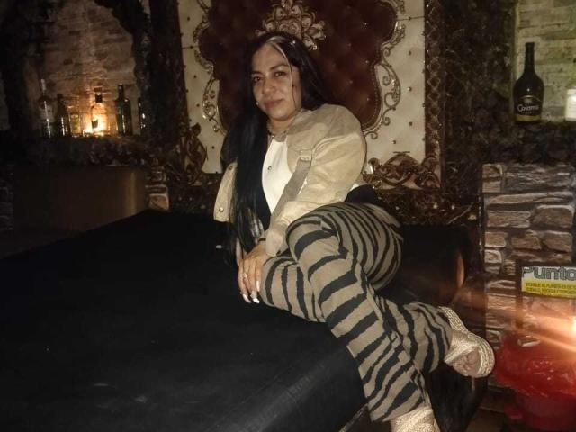 EvaLunaLatina, with medium tits, black hair, tan jacket, white shirt, zebra pants, and tan heels, lounging on a black-covered table in a dark, ornate room.