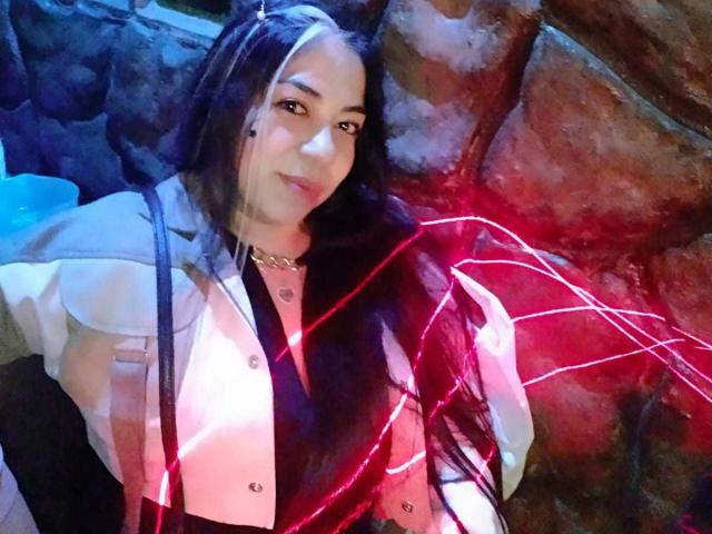 EvaLunaLatina with long black hair, wearing a white jacket, black top, and gold necklace, smiling, red light trails across her face and chest in a dark, rocky setting.