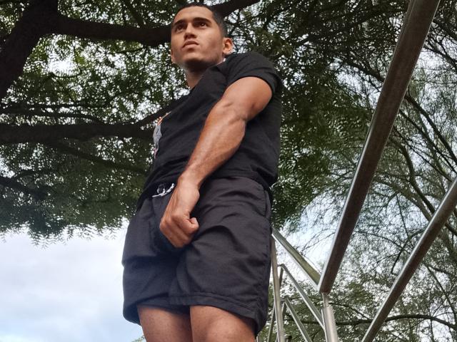 DereckStorm standing tall, muscular arms, black shirt, black shorts, looking up, short dark hair, slight smile, tree branches overhead, metal structure on right, cloudy sky background.