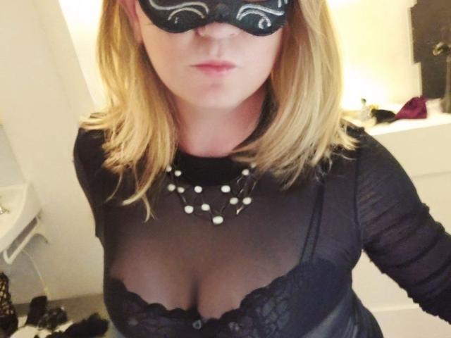 LauralaLouve with blonde hair, black mask, and sheer top showing her tits, wearing a polka dot necklace, in a bathroom. Her face is partially hidden by the mask.
