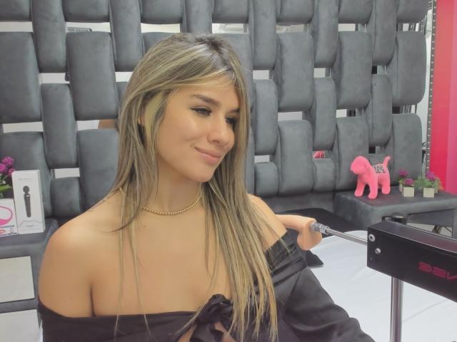 SashaPolet with long blonde hair, tan skin, and small tits, wearing a black off-shoulder top, gold necklace, and subtle makeup, smiling slightly in a salon chair.