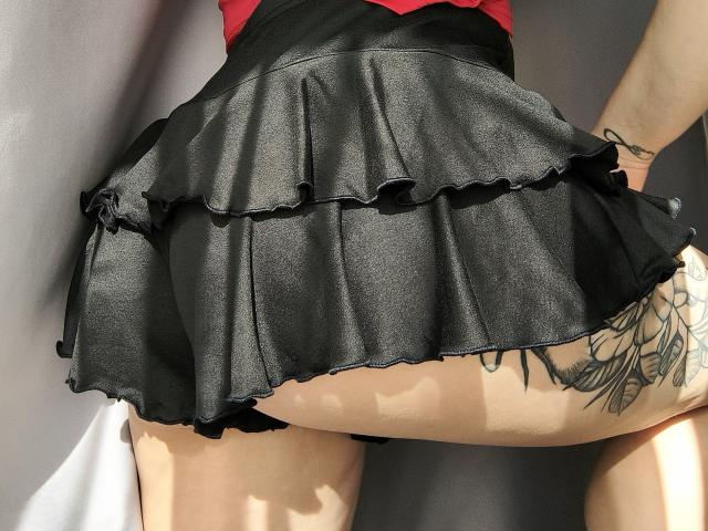 Niferttiti's black, ruffled, mini-skirt is lifted, showing a tattooed thigh and black underwear. Sunlight highlights the skirt's texture and Niferttiti's pale skin.