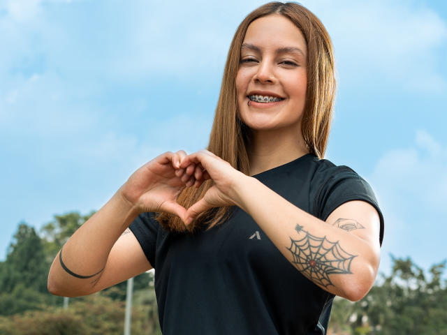 Victoria Ortega, met rechtbruin haar, lachend, gekleed in een zwarte blouse, die een hartvorm maakt met haar handen, met een tatoeage van een spin op haar rechteronderarm, blauwe lucht..