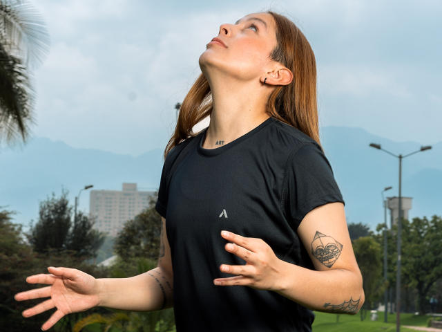 Victoria Ortega, com cabelo castanho, camisa preta, tatuagens no braço, olhando para cima, com as mãos levantadas. Seus seios são pequenos, ela é uma garota atraente. Cenário ao ar livre, céu nublado, árvores..