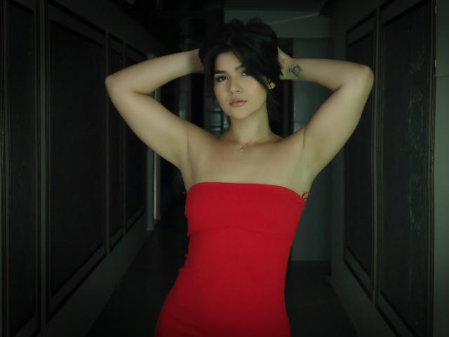 ScarlletZhen, con cabello negro corto, brazos levantados, vestida con un vestido rojo sin tirantes, que deja ver sus pechos y axilas, de pie en un pasillo oscuro, mirando a la cámara..