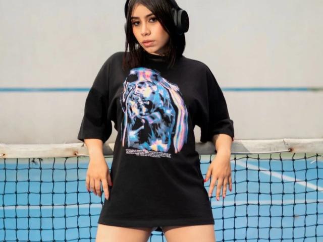 LanaYourDream with black headphones, wearing a black shirt with a colorful tiger graphic, standing in front of a blue tennis court, arms relaxed, looking serious.