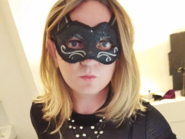 LauralaLouve with blonde hair, wearing a black mask with white swirls, black top with white polka dots, and a serious expression. Her eyes are visible through the mask.