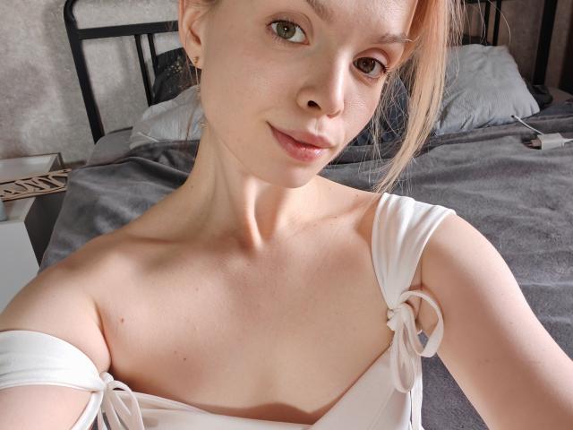 Sinteas with pale skin, blonde hair in a ponytail, small tits, wearing a white off-shoulder top, smiling at the camera, green eyes, soft light, bedroom background.