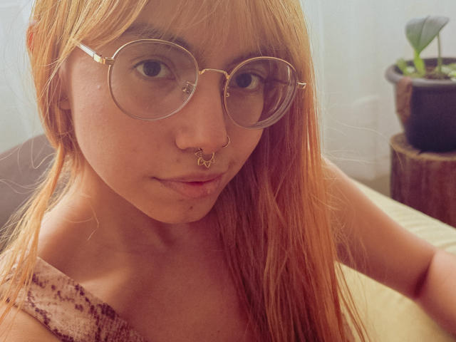 NaturalMariaHun with long orange hair, wearing gold-rimmed glasses, nose ring, and lip piercing, looking at camera, showing bare shoulders, subtle smile, sitting in soft light.