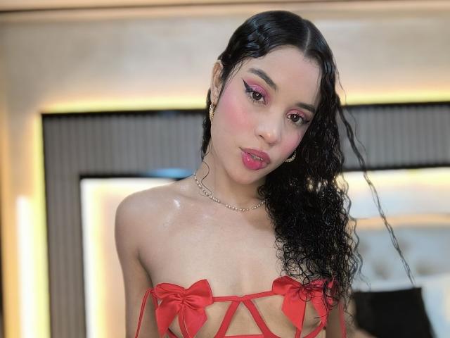 LunaVali with shiny skin, pink eyeshadow, and curly black hair, wearing a red strappy top with small bows, pouting pink lips, and a gold chain necklace.