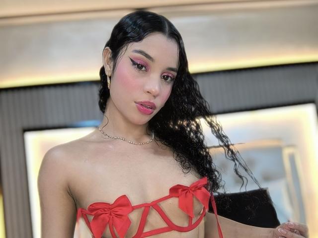 LunaVali with curly black hair, pink eye makeup, red lipstick, wearing a red strappy lingerie top with bows, revealing her small tits, and a gold necklace, looking at camera.